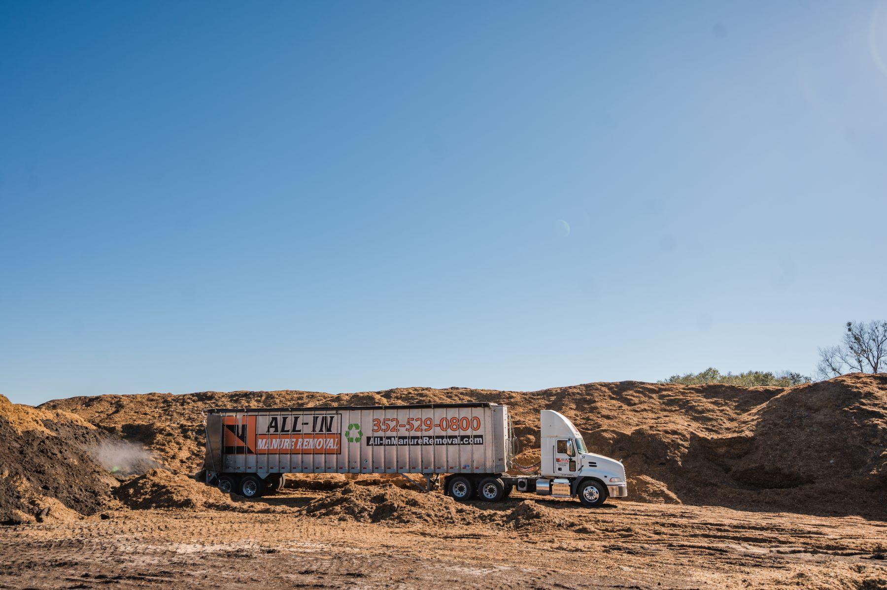 A white semi-truck with a trailer labeled "ALL-IN HAULING REMOVAL" is parked on dirt near large piles of engineered compost under a clear blue sky in Florida.