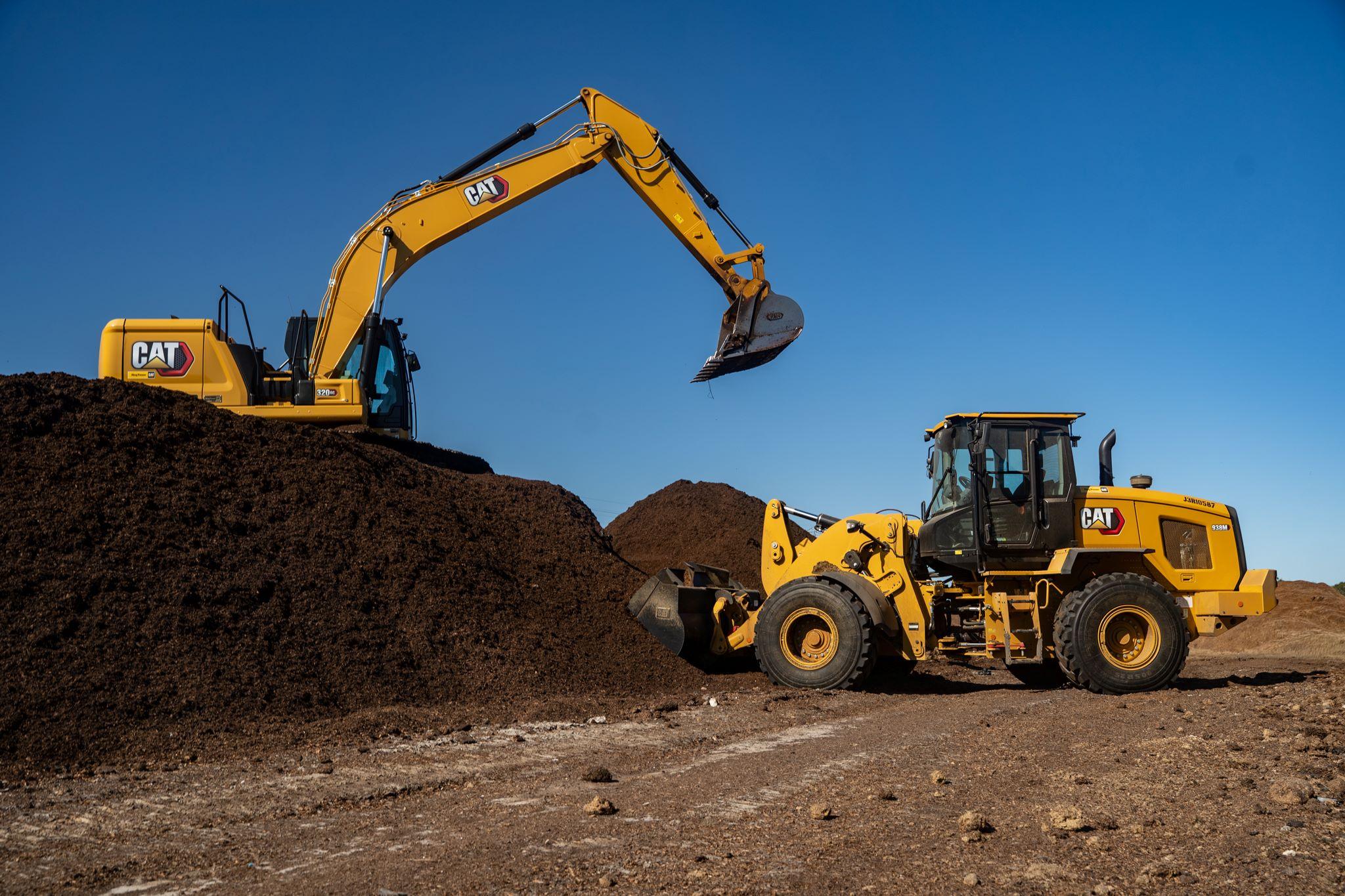 A yellow excavator and wheel loader move large piles of soil or mulch at a construction or work site under a clear blue sky.