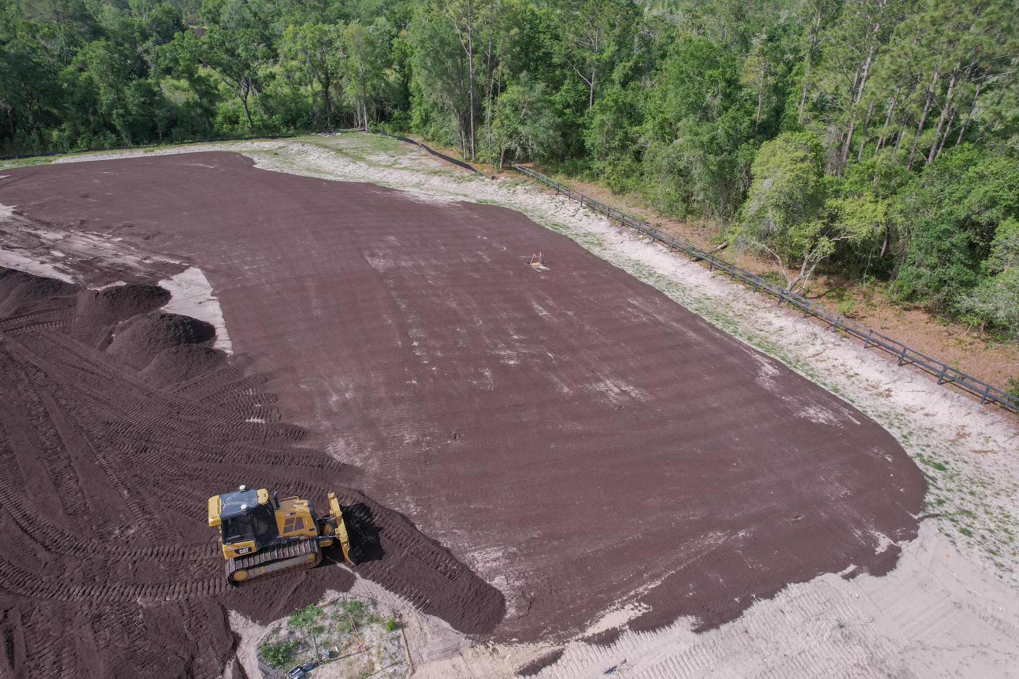 A bulldozer levels a large area of soil near a forested edge, preparing the land for development or landscaping.