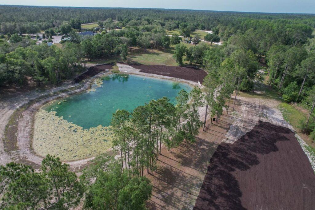 A small pond surrounded by trees and cleared land with patches of dark soil, set in a rural area with houses and forest in the background.