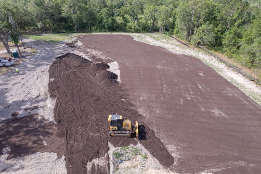 A bulldozer spreads soil across a large, cleared field, with trees surrounding the area and a small vehicle parked to the left.