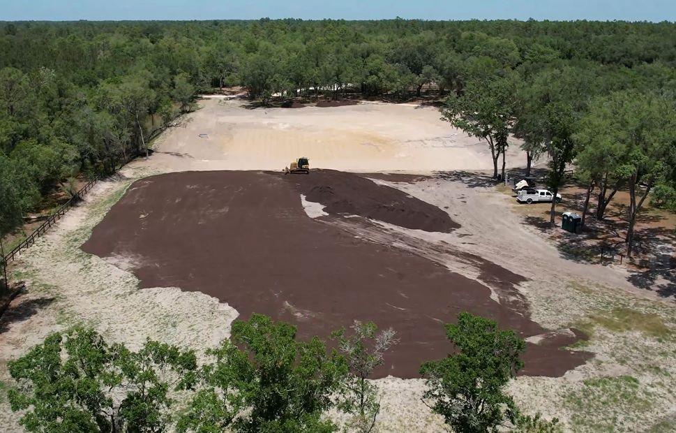 Aerial view of a cleared area in a forest with construction equipment spreading dark soil and vehicles parked nearby.