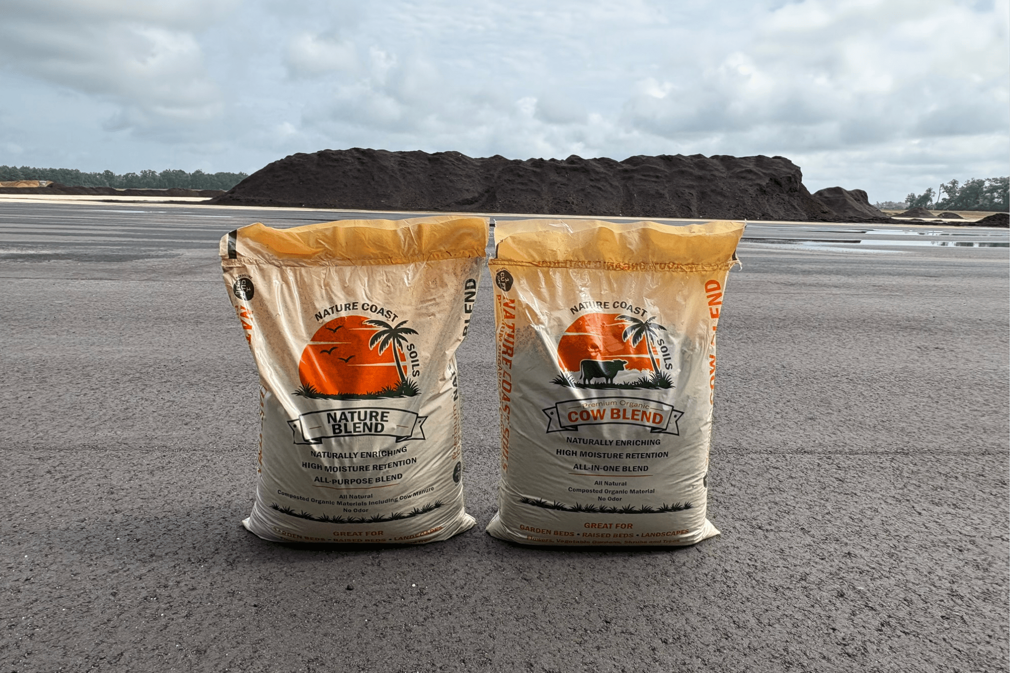 Two large bags of mulch labeled "Nature Blend" and "Cow Blend" stand on asphalt with a large pile of engineered compost Florida and a cloudy sky in the background.