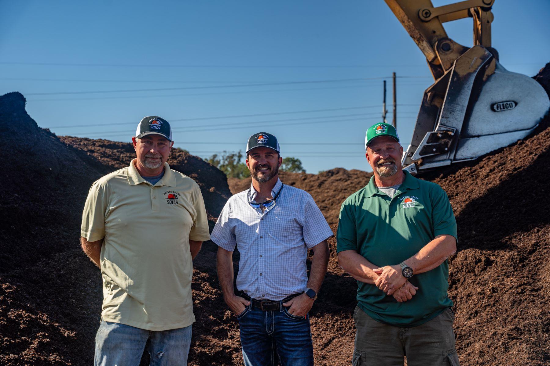 Three men wearing branded shirts and hats stand in front of piles of soil at an organic soil producer in North Florida, with an excavator bucket visible in the background on a sunny day.