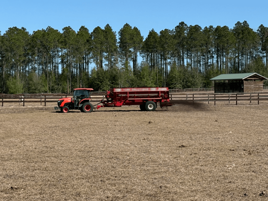 A red tractor pulls a manure spreader across a dry field, with a wooden fence, trees, and a barn visible in the background.
