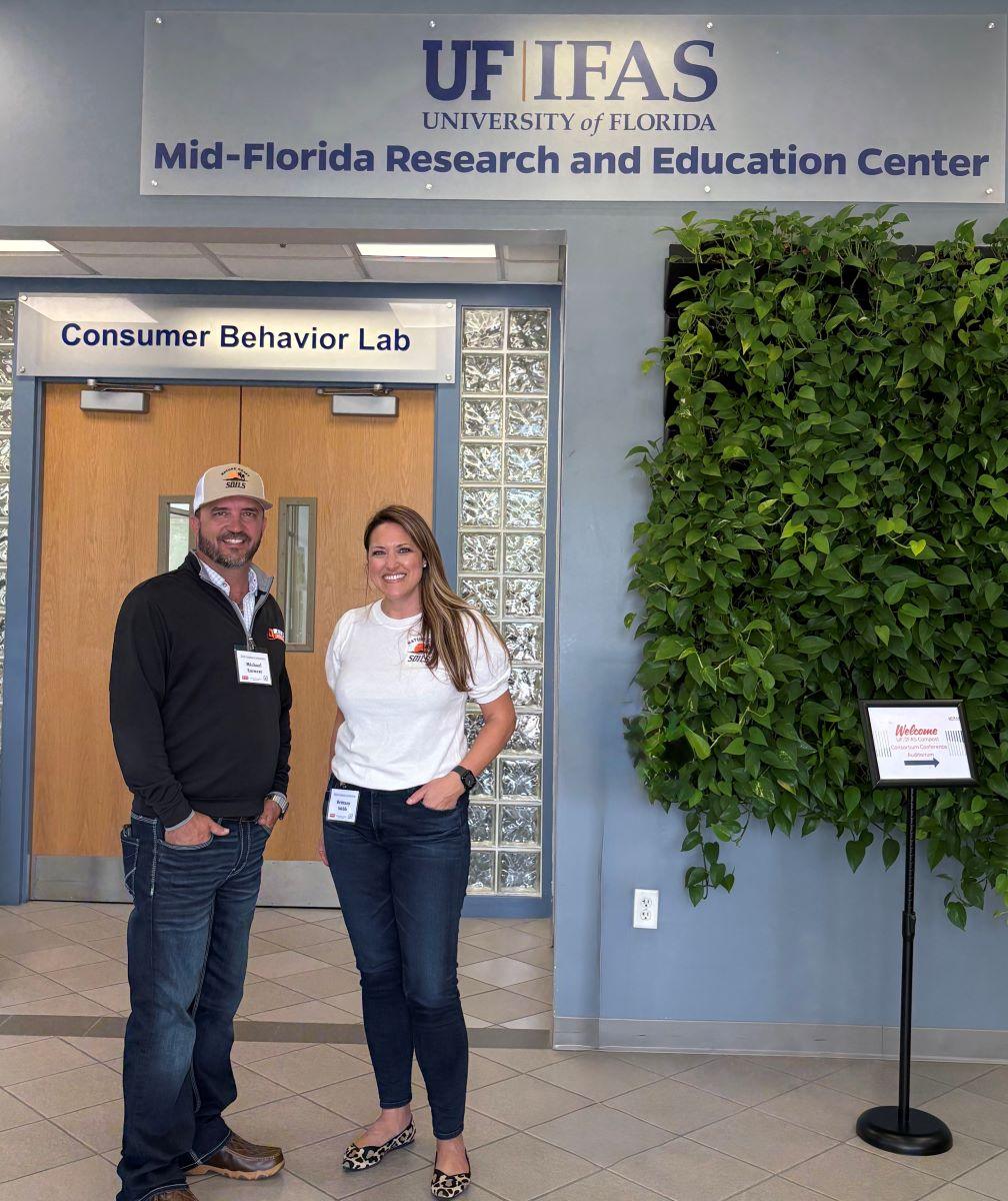Two people stand smiling in front of the Consumer Behavior Lab at the UF IFAS Mid-Florida Research and Education Center.