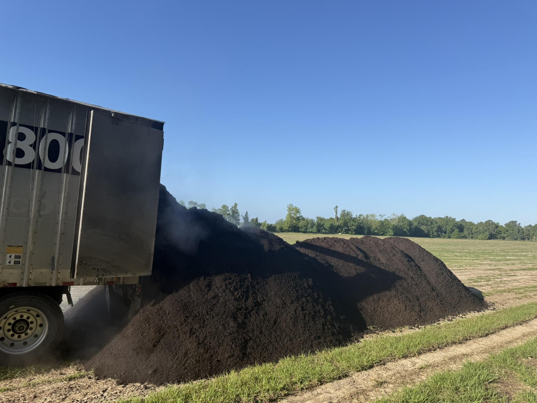 A truck is unloading a large pile of bulk organic soil onto a field, ideal for Florida wholesale needs, with trees and blue sky in the background.