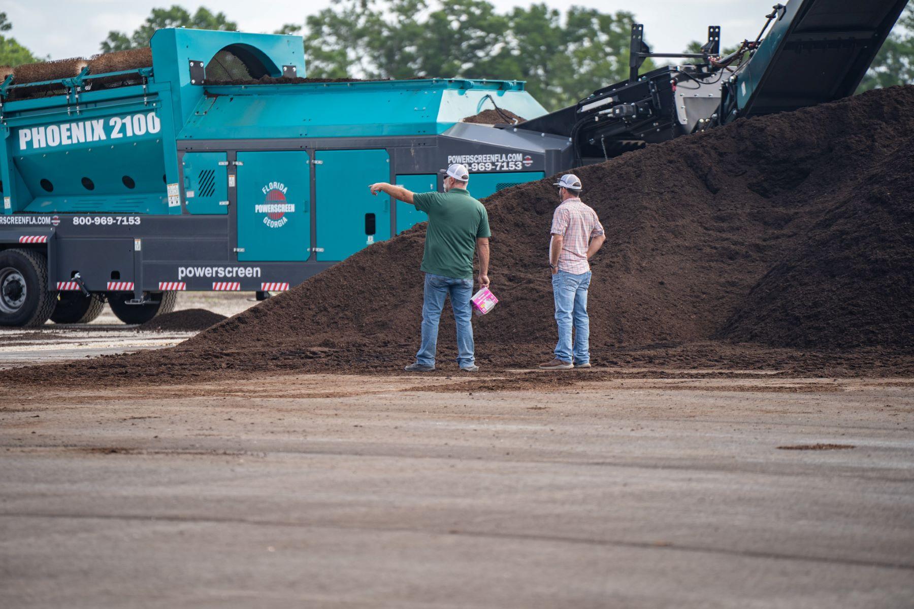 Two men, representing an organic soil producer in North Florida, stand beside a large pile of soil near a blue industrial machine labeled "PHOENIX 2100" and "Powerscreen," with one man pointing towards the equipment.