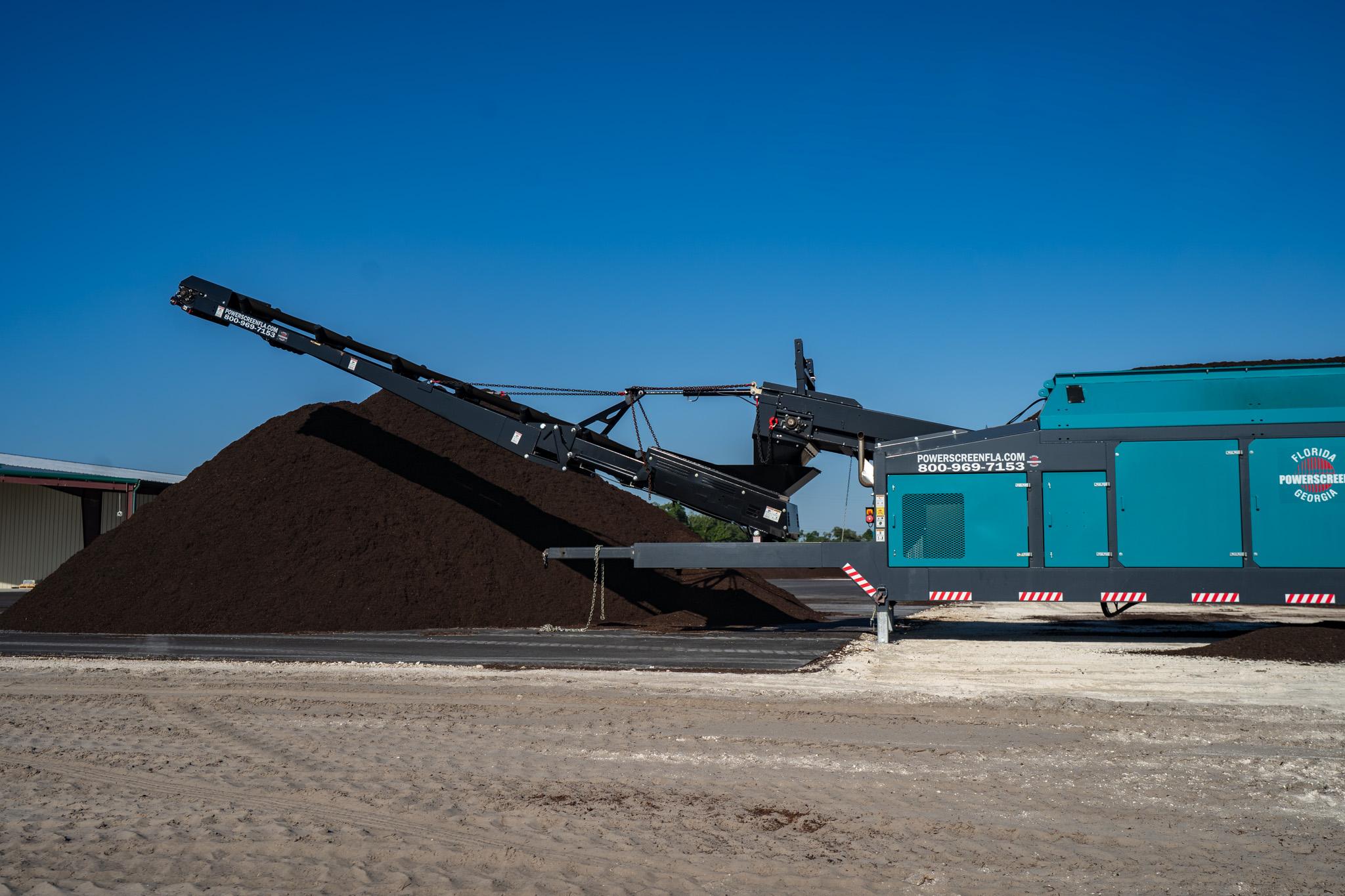 A conveyor belt machine deposits a large pile of dark soil or compost onto the ground at an SOPF registered composting facility in Florida, set outdoors under a clear blue sky.