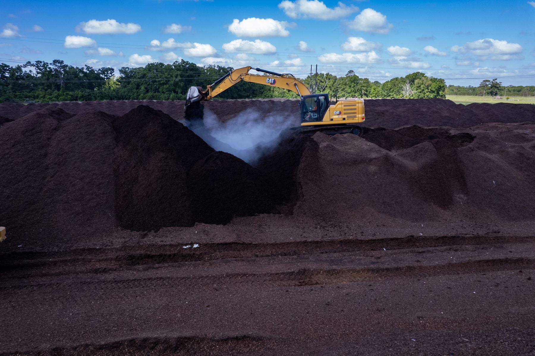 A yellow excavator moves bulk organic soil in a large outdoor pile, with dust rising from the soil under a partly cloudy Florida sky.
