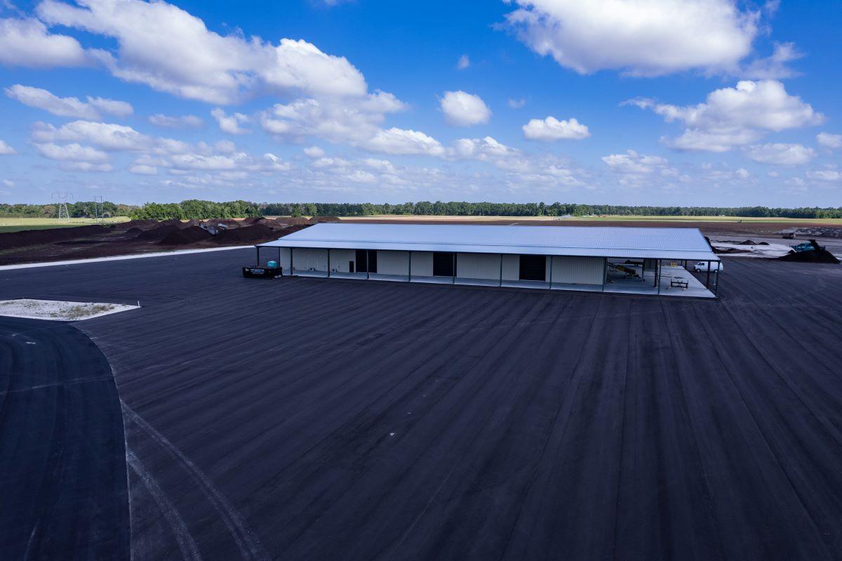 A large, single-story metal warehouse stands on a newly paved lot under a blue sky with scattered clouds, storing bulk organic soil for Florida wholesale. Open fields and trees stretch in the background.
