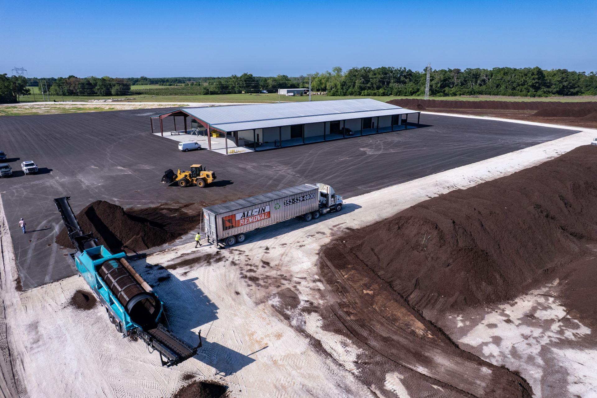 A large industrial facility with soil piles, a conveyor machine, trucks, and workers on a paved lot beside a warehouse building under a clear sky, operated by an organic soil producer in North Florida.