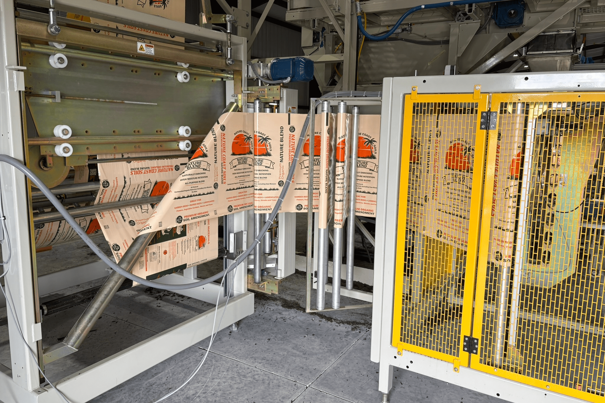 Automated packaging machine feeds printed brown paper bags featuring orange graphics and text through rollers and metal guides, with yellow safety barriers on the right.