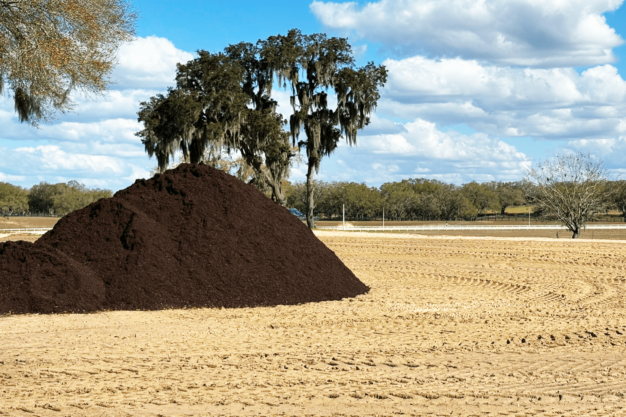 A large pile of all purpose organic soil amendment sits on a sandy field with tire tracks, trees in the background, and a partly cloudy Florida sky overhead.