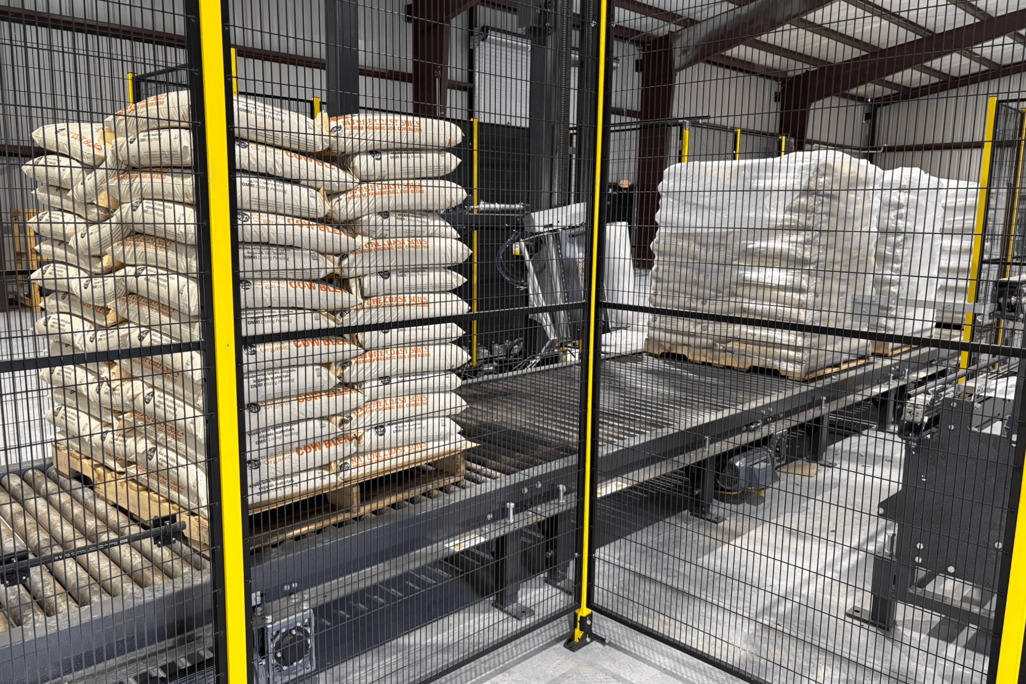 Stacks of bagged goods on pallets are positioned on a conveyor system inside an industrial warehouse, enclosed by a metal safety fence.