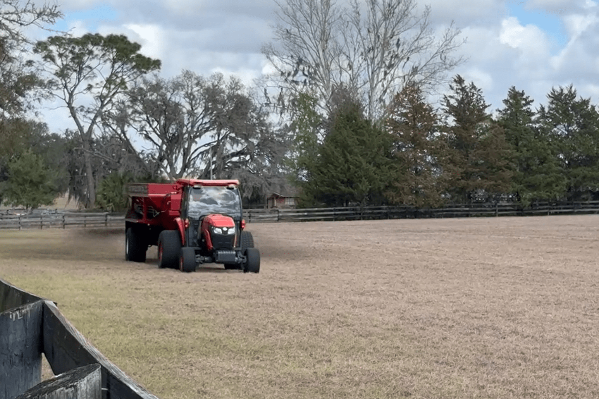 A red tractor spreads material on a dry, grassy field bordered by a wooden fence and trees under a partly cloudy sky.