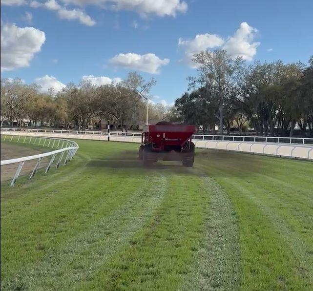 A red tractor spreads material on a grassy horse racing track under a partly cloudy sky, surrounded by trees and white railings.