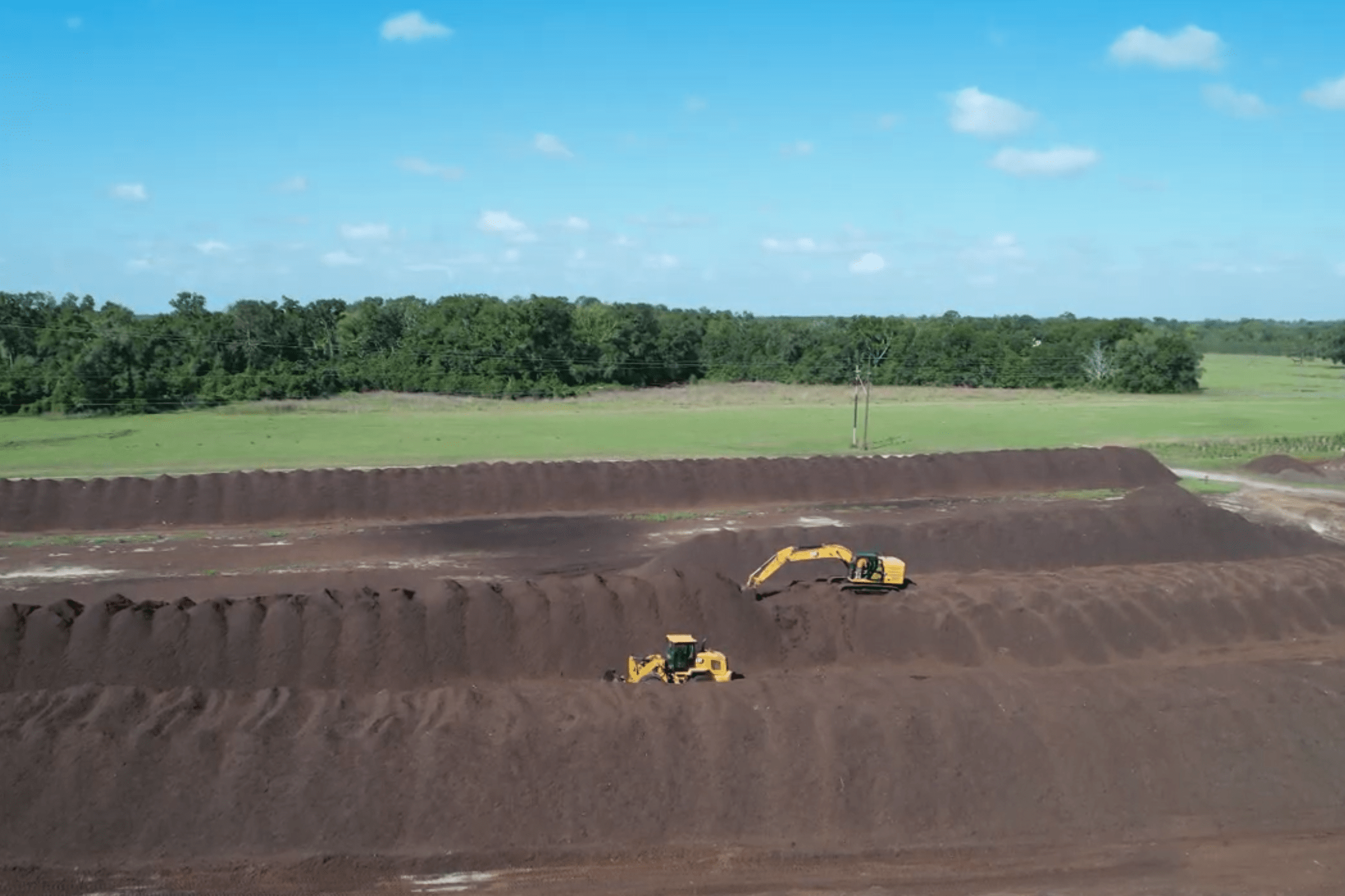 Two excavators move soil on large mounds at an open field under a blue sky, with trees and grassy areas in the background.