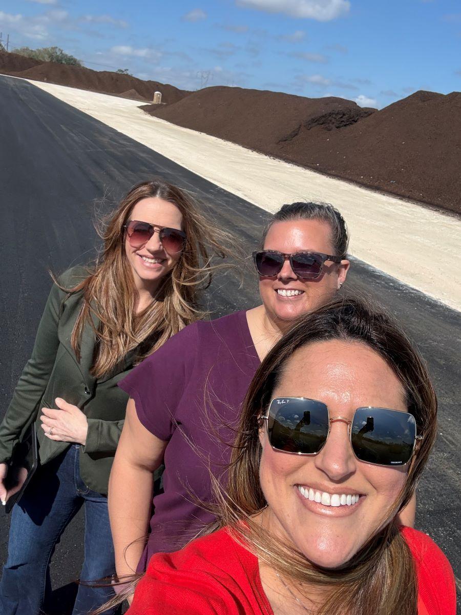 Three women wearing sunglasses smile for a selfie outdoors on a paved road with large piles of soil in the background under a blue sky.