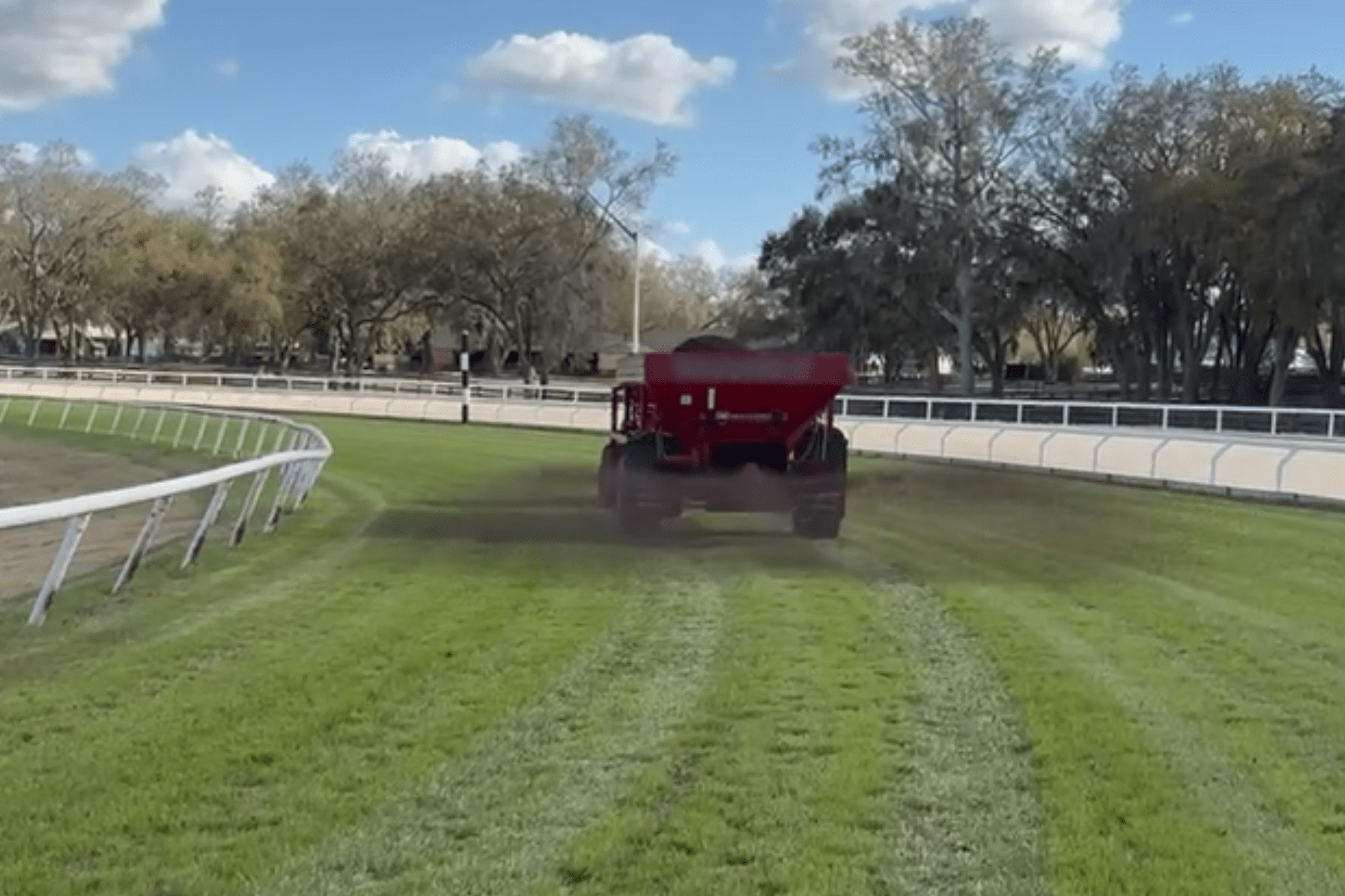 A red agricultural vehicle spreads material on a grassy racetrack, leaving visible tracks; trees and a white fence line the background under a partly cloudy sky.
